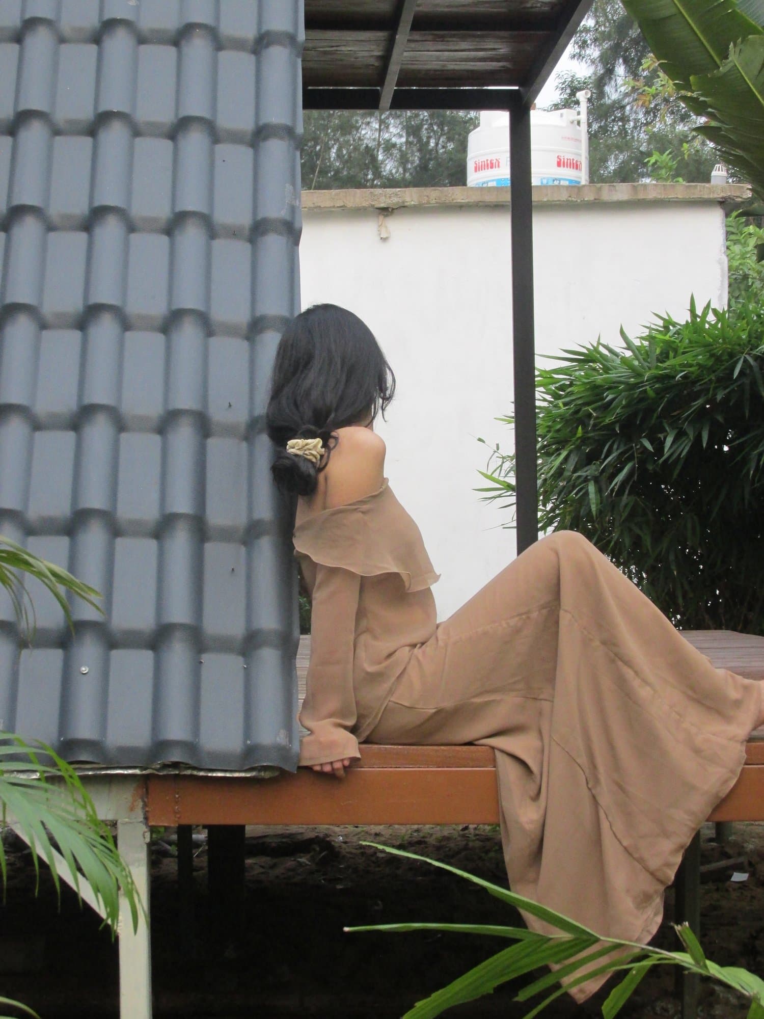 A woman in a camel dress seated in profile on a bench, looking out over a green garden.